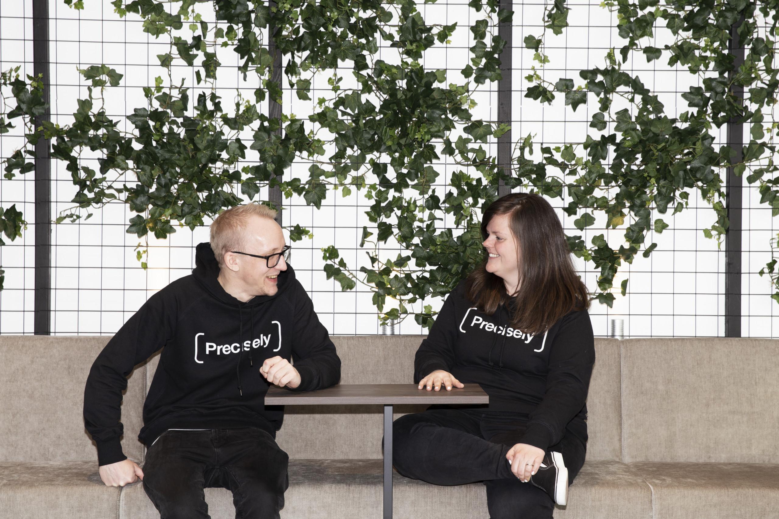 Two happy people sitting in front of a plant wall wearing black hoodies with Precisely logos