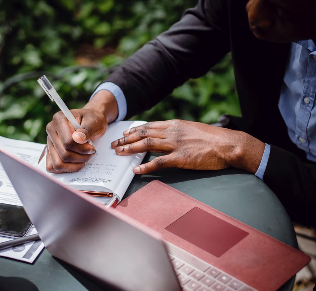 hands writing in notebook next to laptop